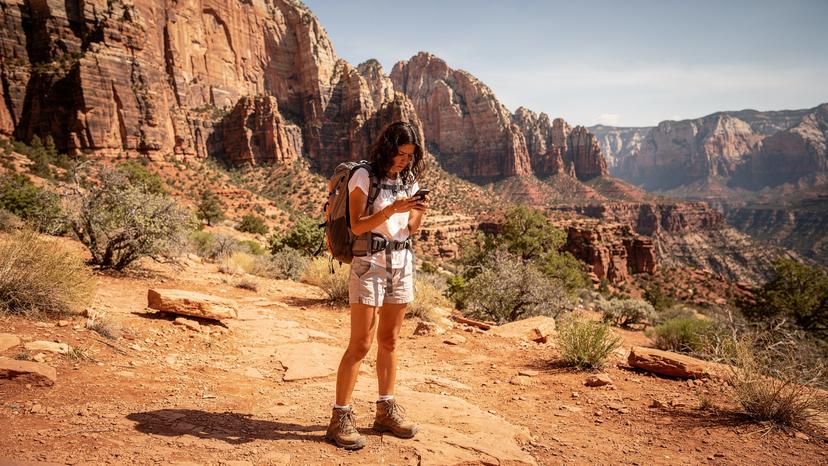 Una donna italiana osserva con aria preoccupata il suo smartphone con "Nessun Servizio" sullo schermo mentre si trova in un'area remota del Parco Nazionale di Zion, con maestose formazioni rocciose rosse in sottofondo.
