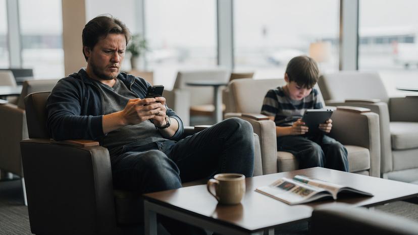 En förälder ser irriterad ut på sin telefon medans barnet i bakgrunden spelar på en surfplatta. Scenen är i en flygplatslounge eller hotellobby.