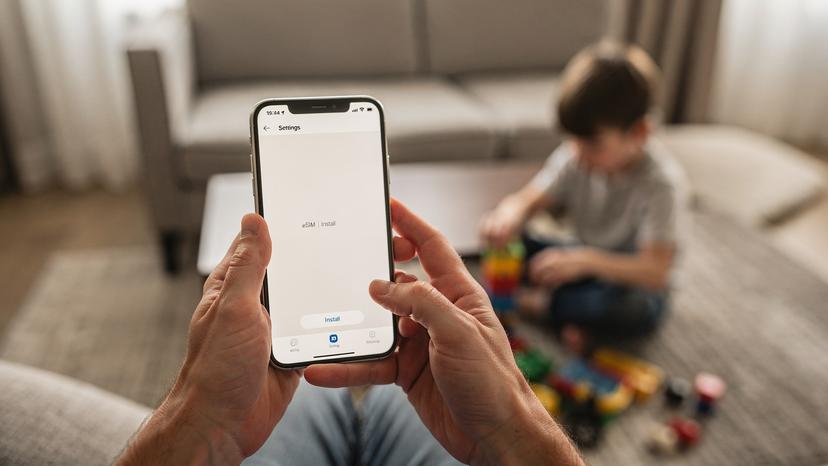 En förälder håller sin smartphone och visar en skärm med eSIM-installationsinstruktioner. Ett barn leker i bakgrunden. Scenen utspelar sig i ett lugnt rum.
