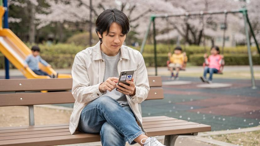 日本の公園で、子供たちが楽しそうに遊んでいる間に、親がスマートフォンを使い、CellesimとトリファのeSIMアプリの比較画面を真剣に見ている。