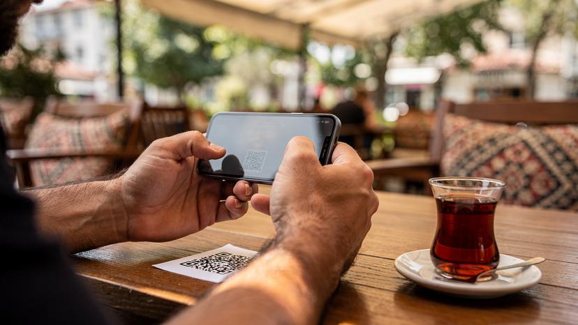 Мъж сканира QR код от разпечатан документ с камерата на своя iPhone 18, докато седи на маса в традиционно турско кафене в квартал Доганджилар, Истанбул, с чаша чай до него.