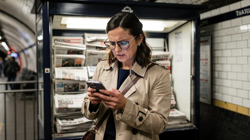Una donna con occhiali legge lo schermo del suo smartphone di fronte a un chiosco di giornali tradizionale in una stazione della metropolitana di Londra, visibilmente in difficoltà con la connessione.