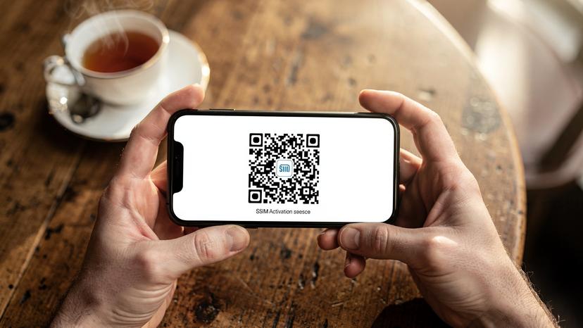 Mani che tengono uno smartphone con un QR code di attivazione eSIM sullo schermo, con uno sfondo sfocato di un tavolino da caffè e una tazza di tè inglese.