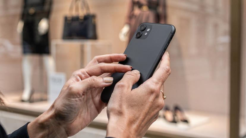 Un primo piano delle mani di una donna italiana mentre confronta prezzi e recensioni di prodotti di moda sul suo smartphone, con lo sfondo sfocato di una vetrina di un negozio di lusso a Boulevard Haussmann, Parigi.